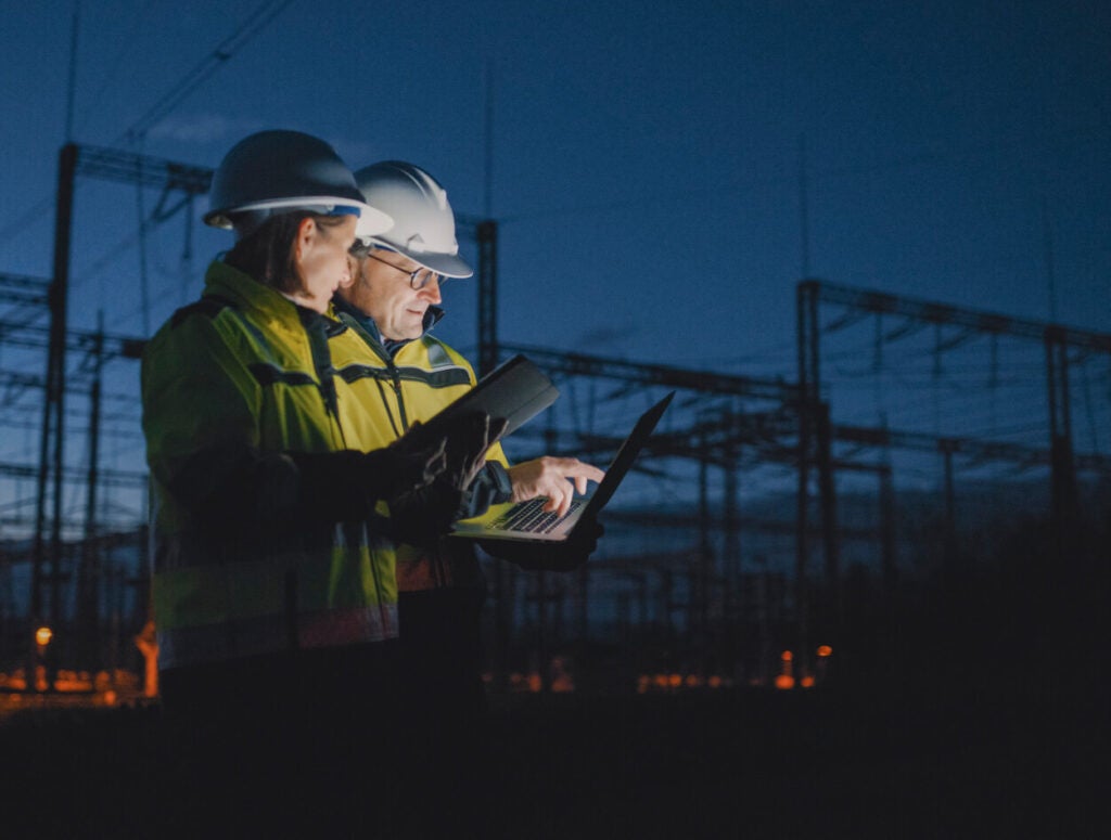 Two people wearing yellow jackets and hard hats working off laptops.