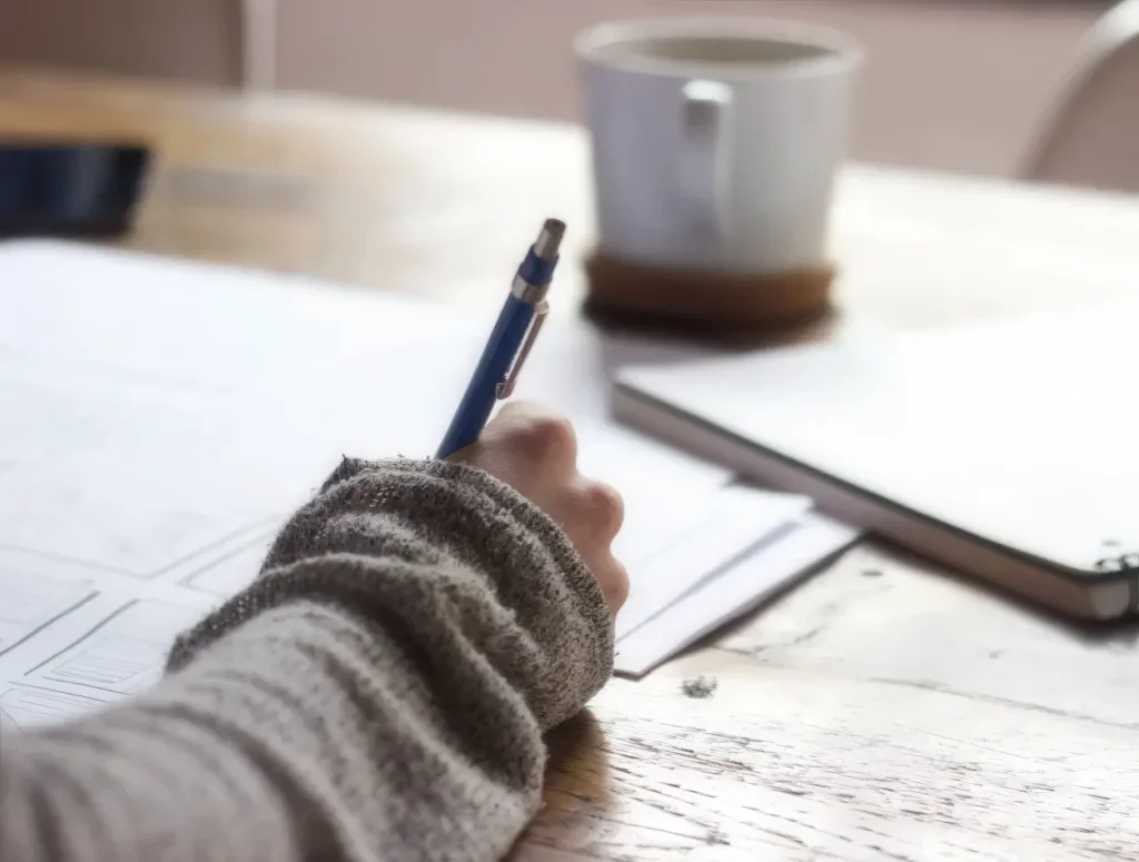 Person, die an einem Schreibtisch mit Stift und Papier Notizen macht.