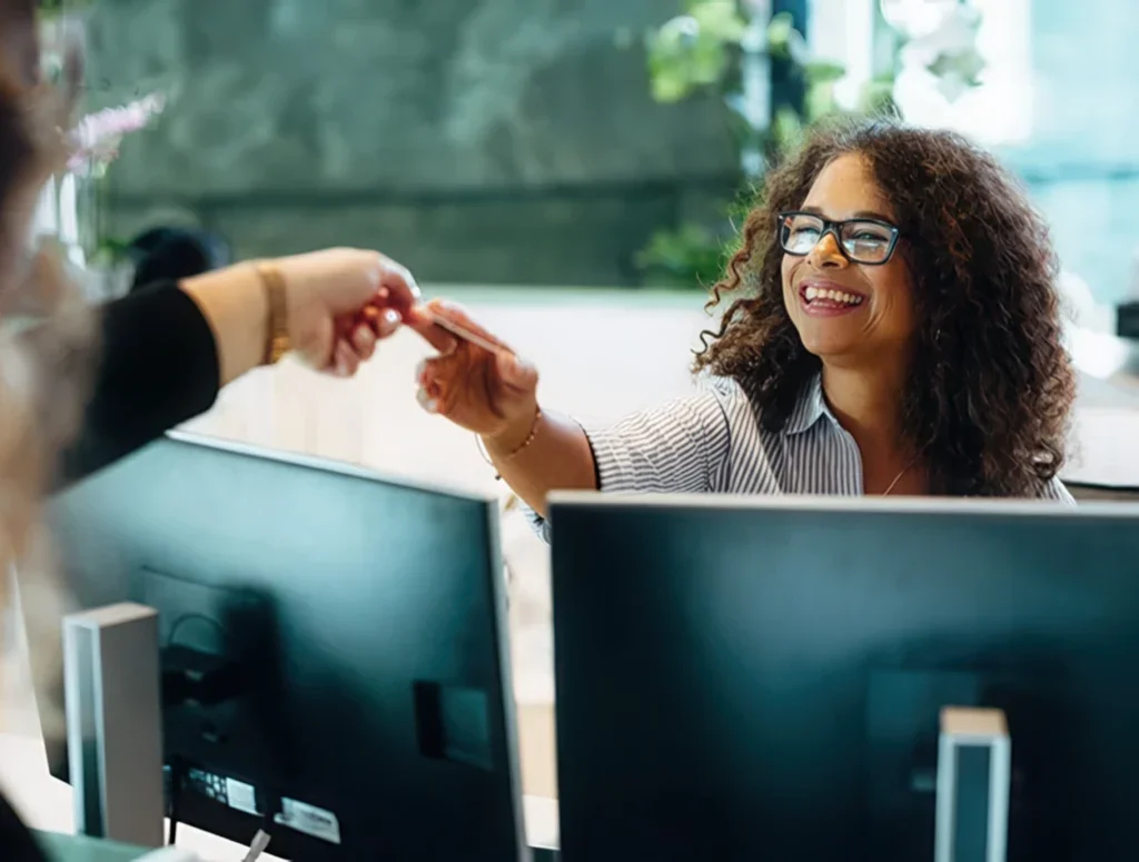 Femme souriante, interagissant avec un autre employé sur un double écran.