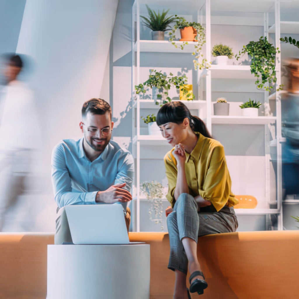 Two people having a conversation over a laptop computer in a casual business office setting.